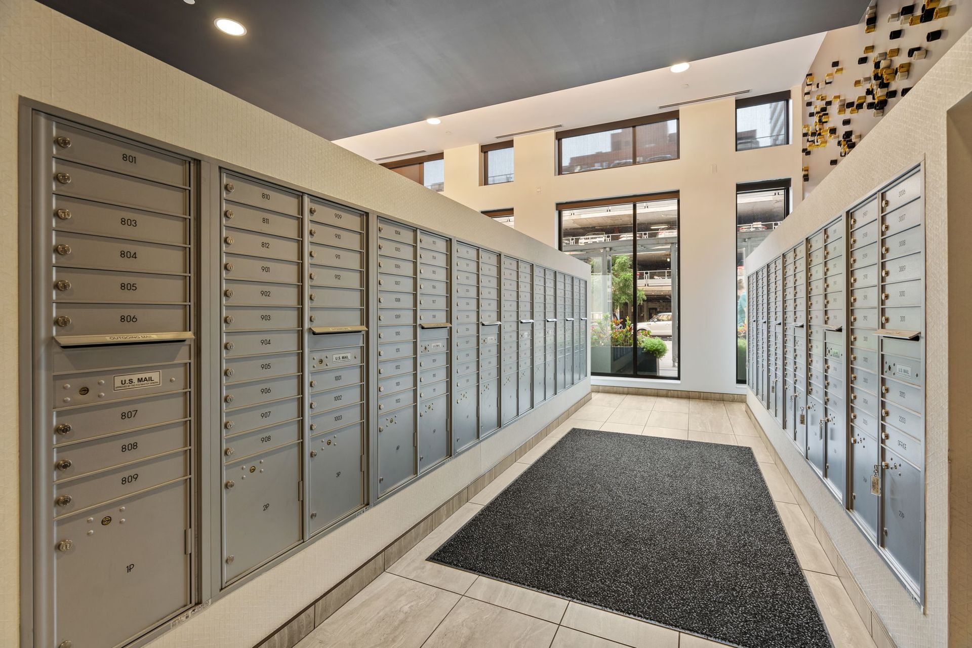 Rows of mailboxes in a bright apartment lobby. A doormat is in the center.