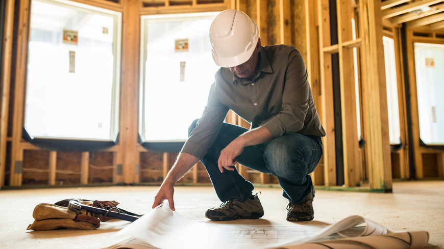 Construction worker kneeling over blueprints in a framed house interior, measuring plans on the floor.
