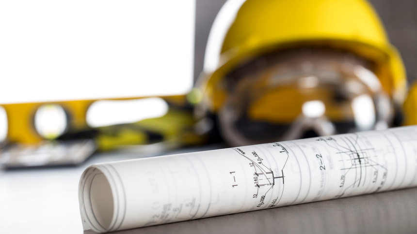 Rolled blueprints with a yellow hard hat and measuring tape on a table
