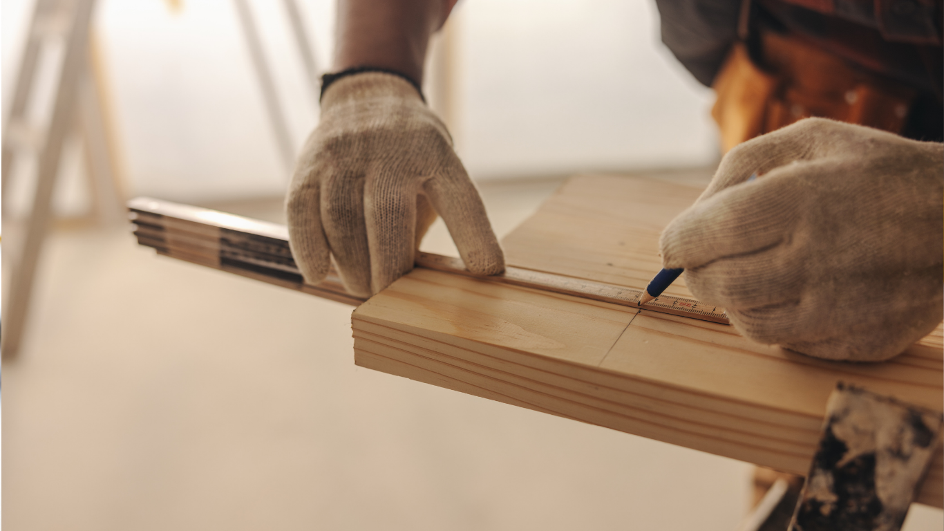 Gloved hands measuring a wooden board with a pencil in a workshop-like setting.