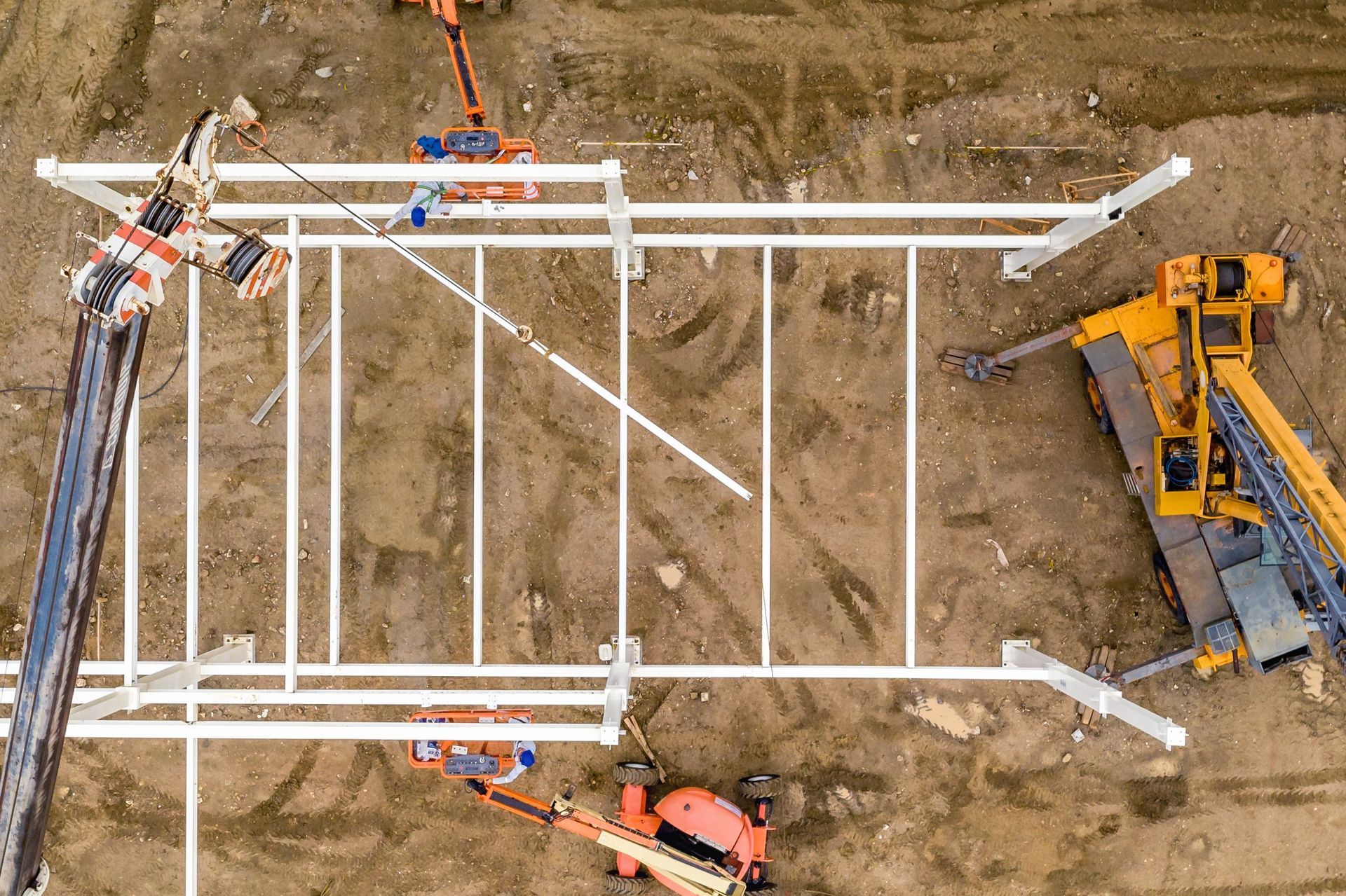 Overhead view of workers assembling a white metal frame beside a yellow excavator on a dirt construction site