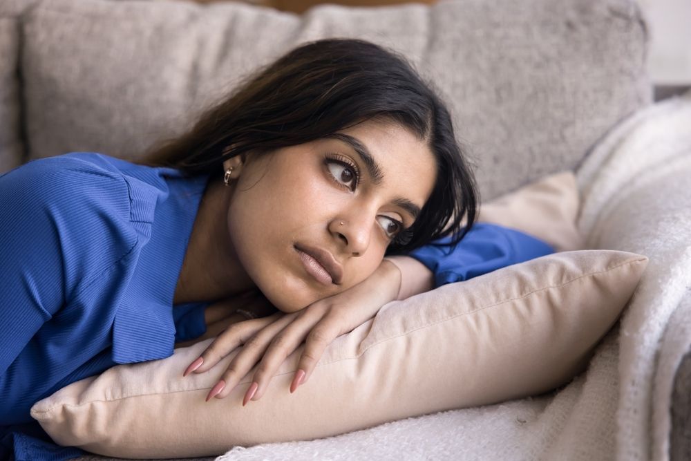 Woman resting on a couch, looking pensive. She is wearing a blue shirt and resting her head on a beige pillow.
