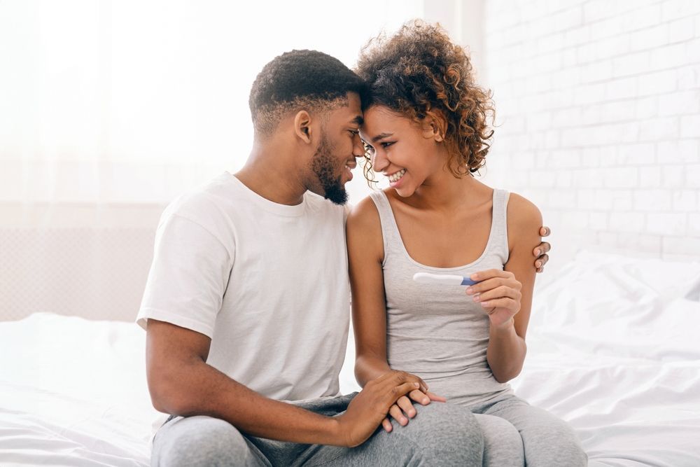 Couple in bed, smiling and looking at a positive pregnancy test; holding hands.