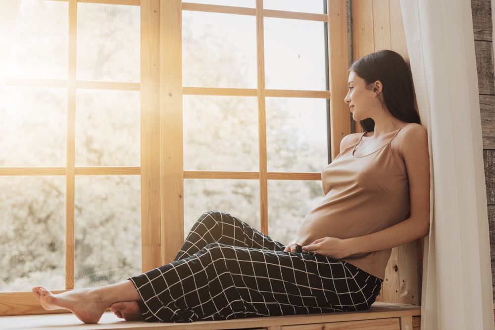 Pregnant woman sitting by a sunlit window, resting with hands on her belly.