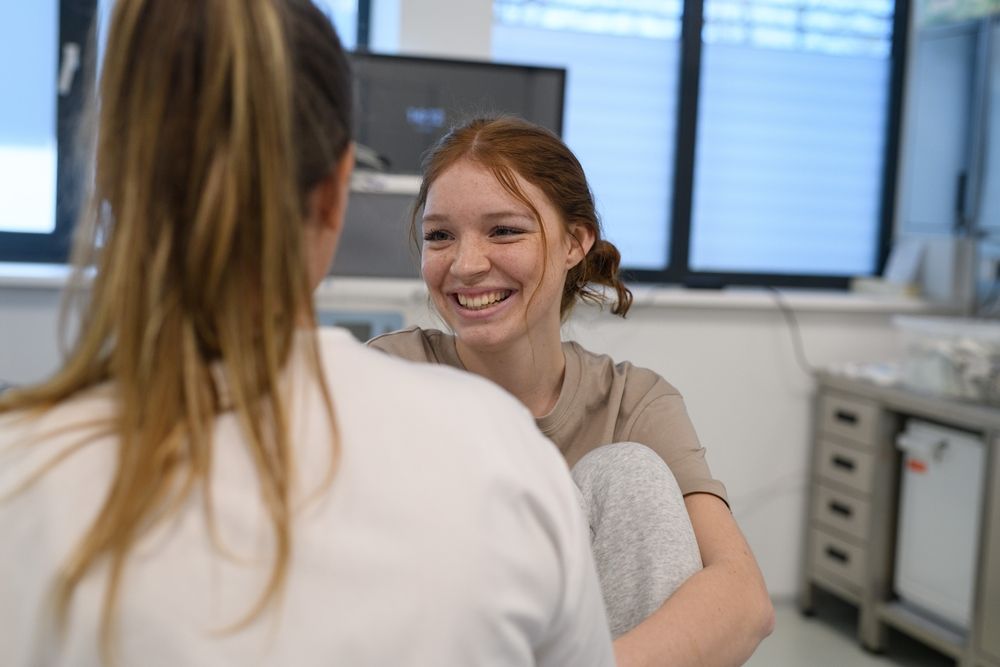 Two women smiling and talking in a well-lit room. One with red hair smiles broadly while the other's back is to the camera.
