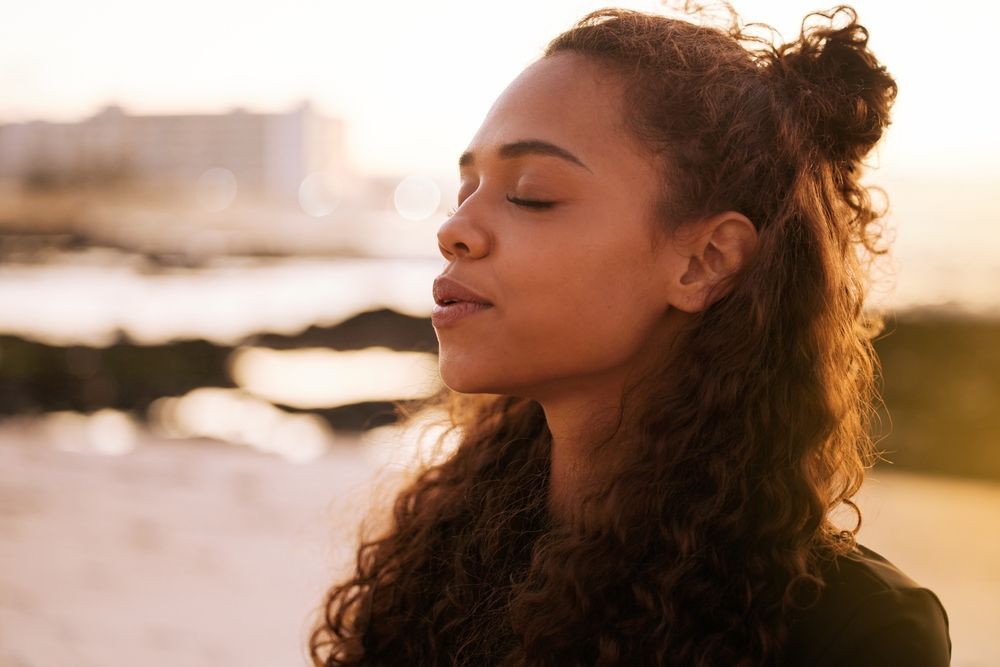Woman with eyes closed, enjoying sunlight near a beach, hair up in a bun.
