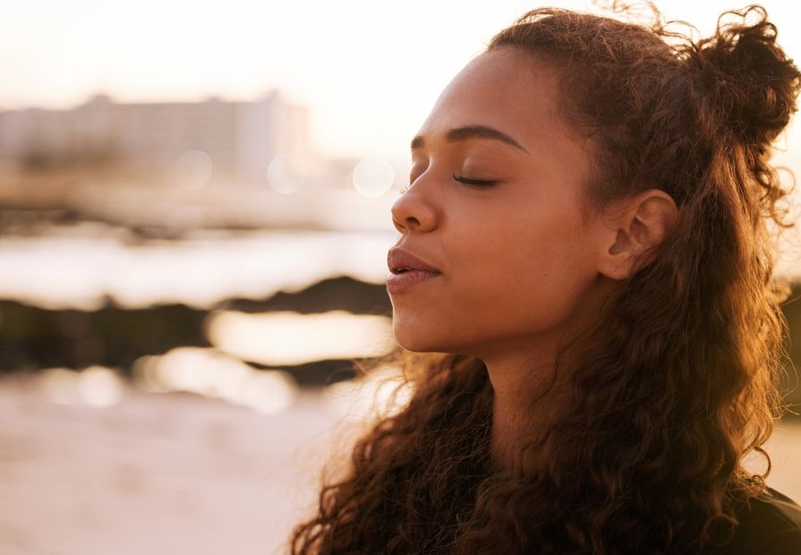 Woman with curly hair closes her eyes, breathing deeply. Outdoors with blurred background of a body of water.