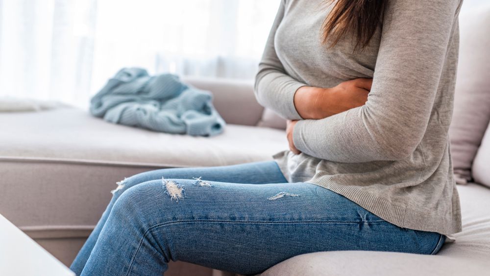 Person sitting on a couch, holding their stomach in apparent pain, with a blanket in the background