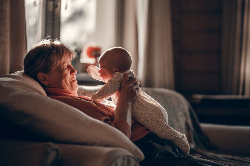 Grandmother holding baby, lit by sunlight, indoors. Warm tones, smiles, and a tender moment.