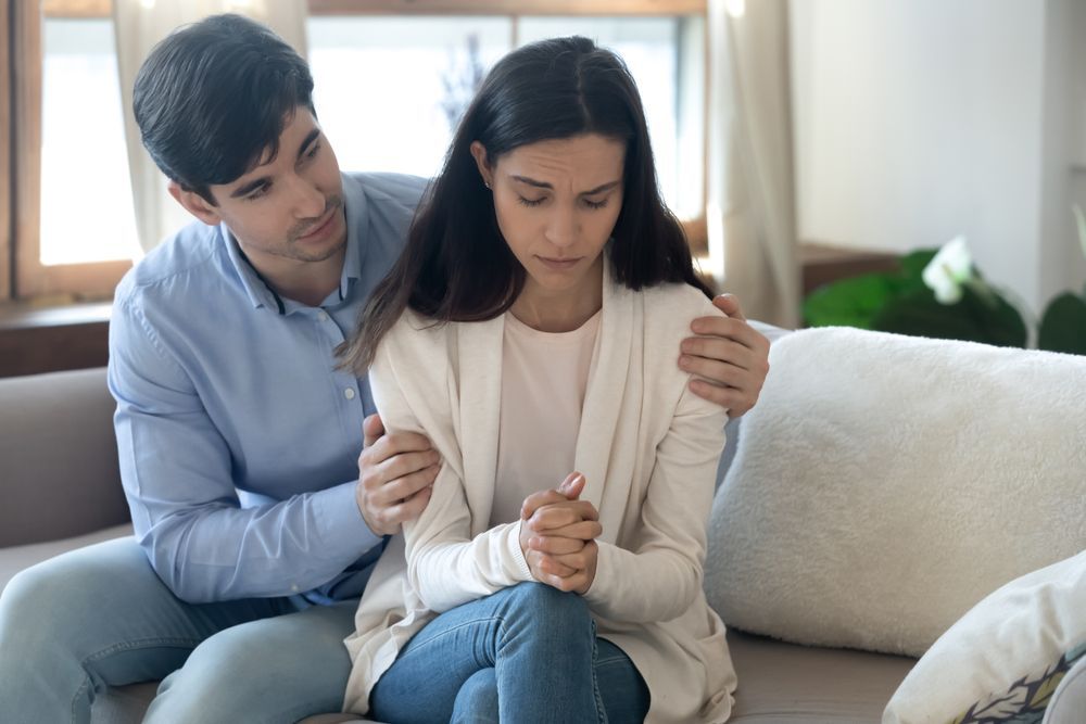 A couple sits on a couch; the man comforts the woman as she looks down sadly.