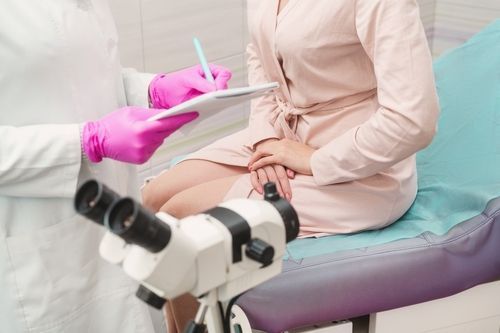 A doctor in pink gloves takes notes while a patient sits on an examination table. A colposcope is in the foreground.