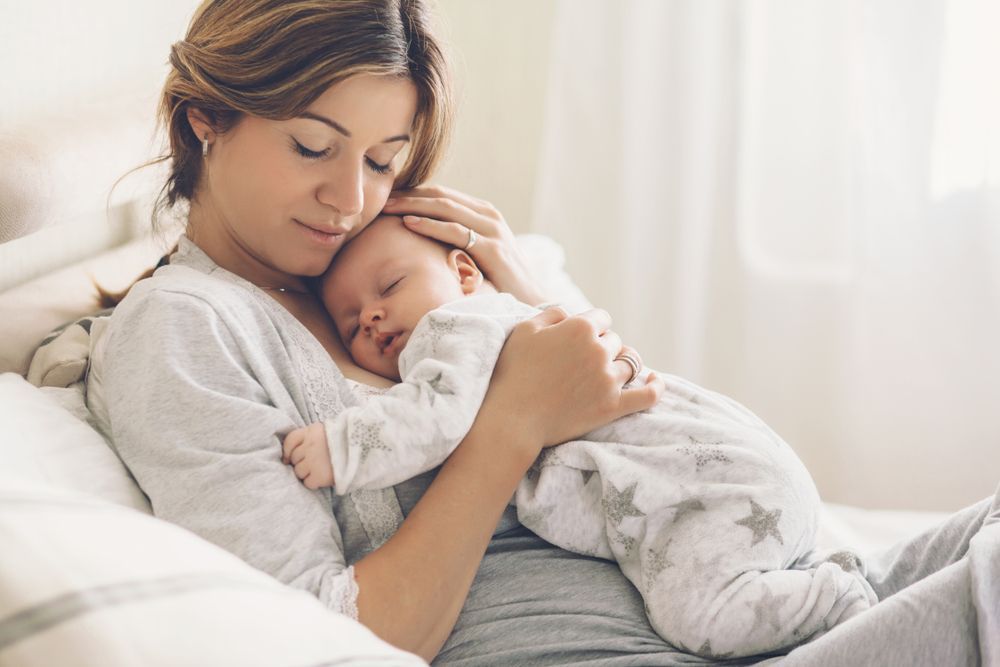 Woman in gray pajamas holding a sleeping baby, both on a bed, serene expression.