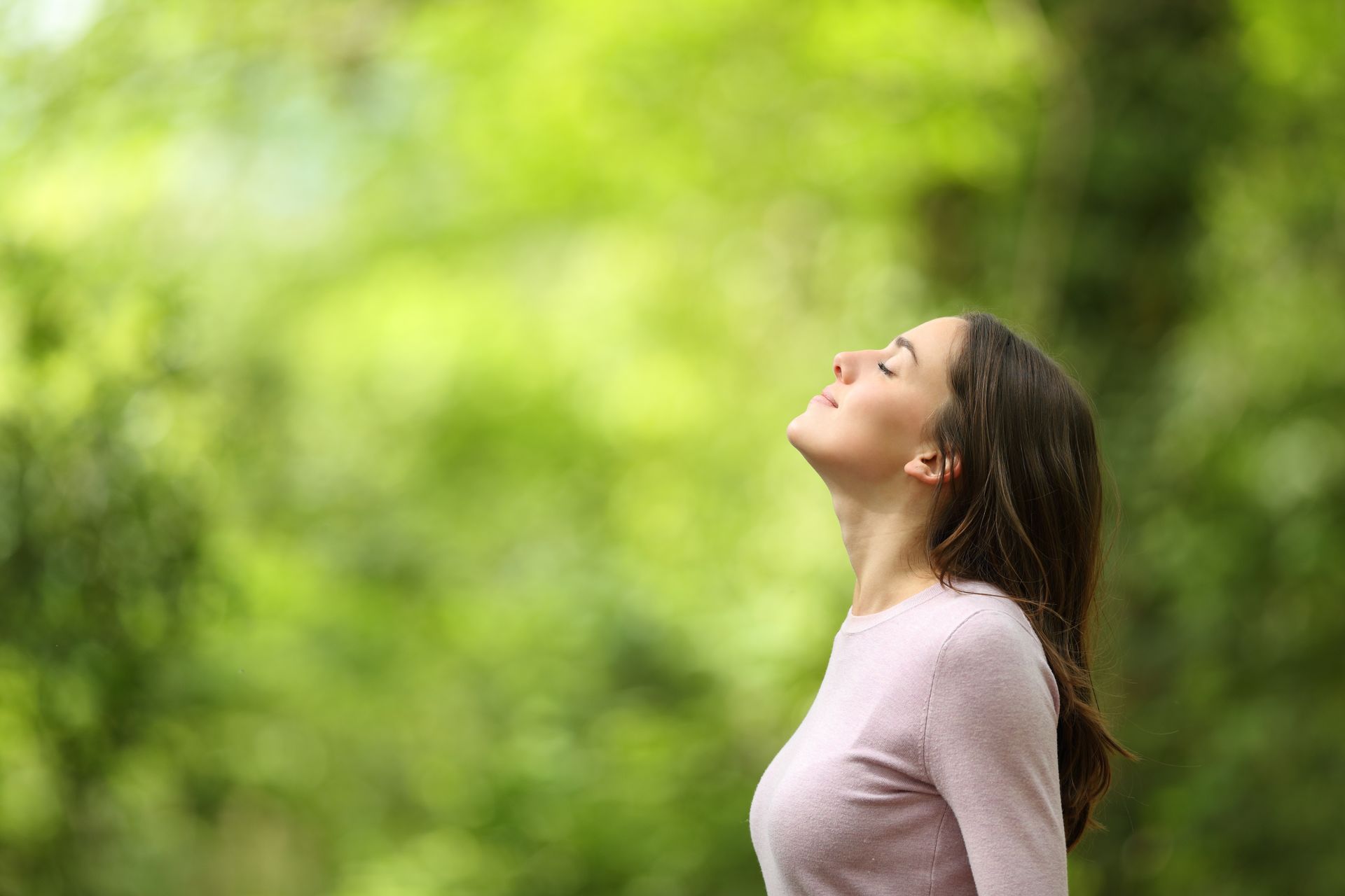 Woman with eyes closed, enjoying fresh air outdoors; green foliage in background.