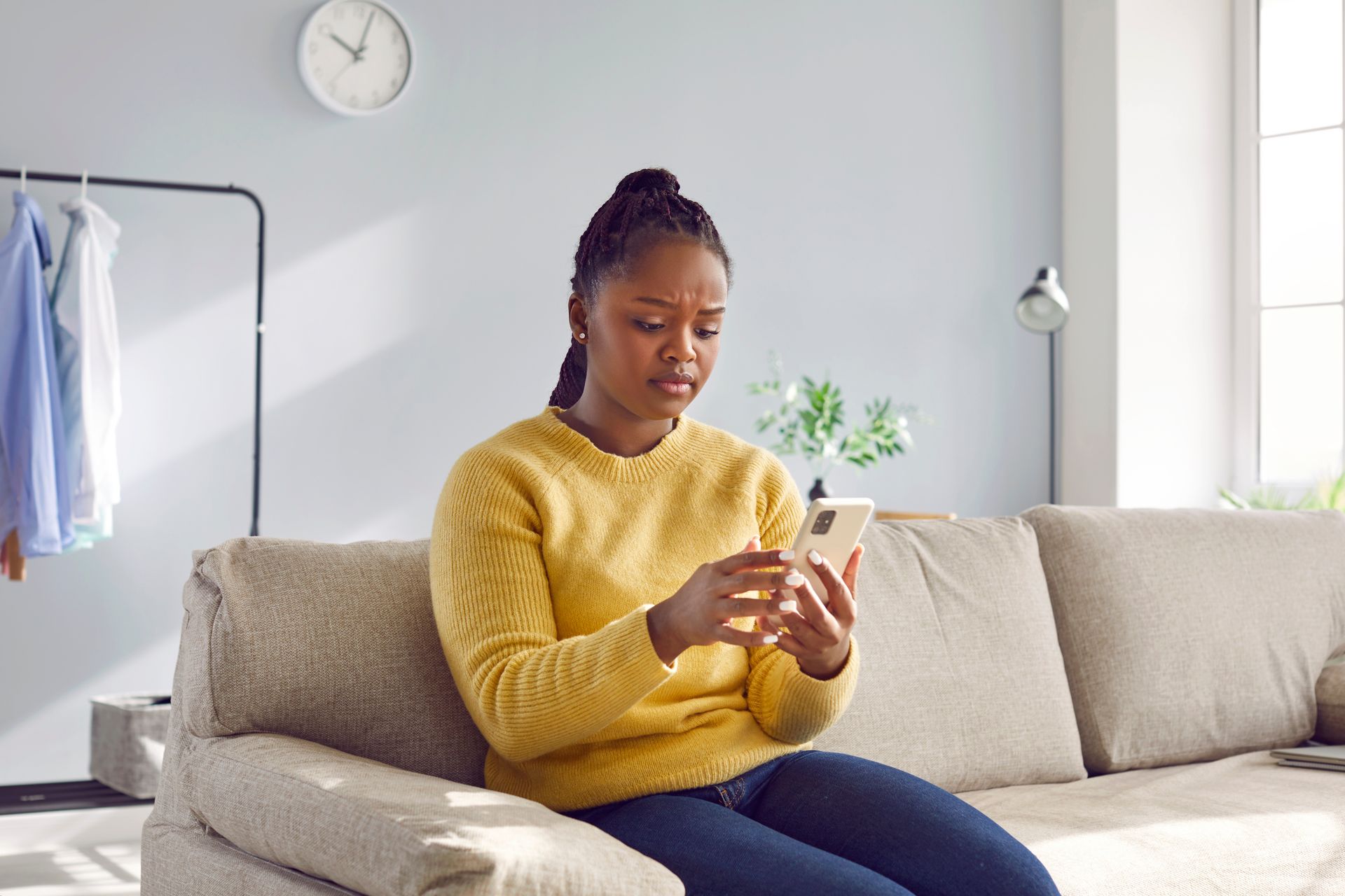 Black woman with a concerned expression, sitting on a couch, looking at her phone in a living room.