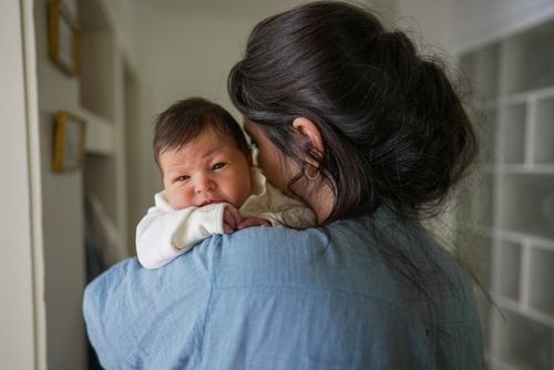 Woman in blue shirt holding a baby over her shoulder; baby gazes at camera.