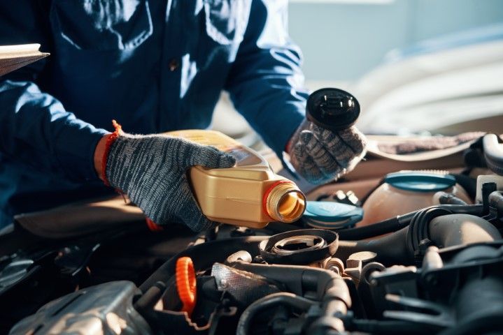 Mechanic pouring oil into a car engine in a garage, with gloved hands and open hood.
