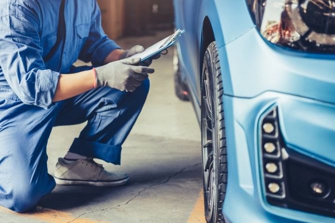 Mechanic in blue coveralls inspecting a blue car’s front wheel with a tool in a garage