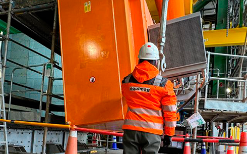 Un trabajador con traje de seguridad naranja y casco se encuentra junto a una gran maquinaria industrial naranja en una obra de construcción.