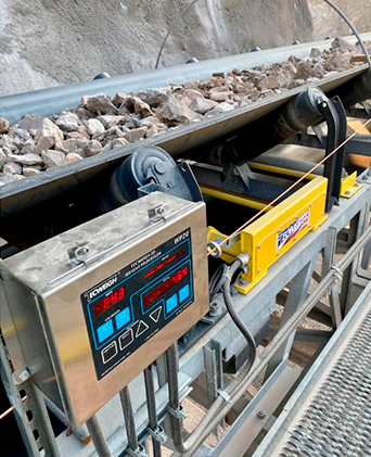 Transportador industrial con rocas y un panel de control digital en una instalación de maquinaria metálica.