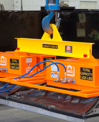 Abrazadera de elevación industrial naranja con gancho azul y mangueras negras, sobre un palé en un almacén.
