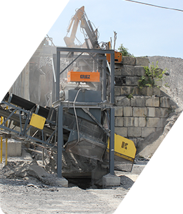 Excavadora triturando hormigón en un solar de demolición lleno de escombros detrás de un muro de piedra.