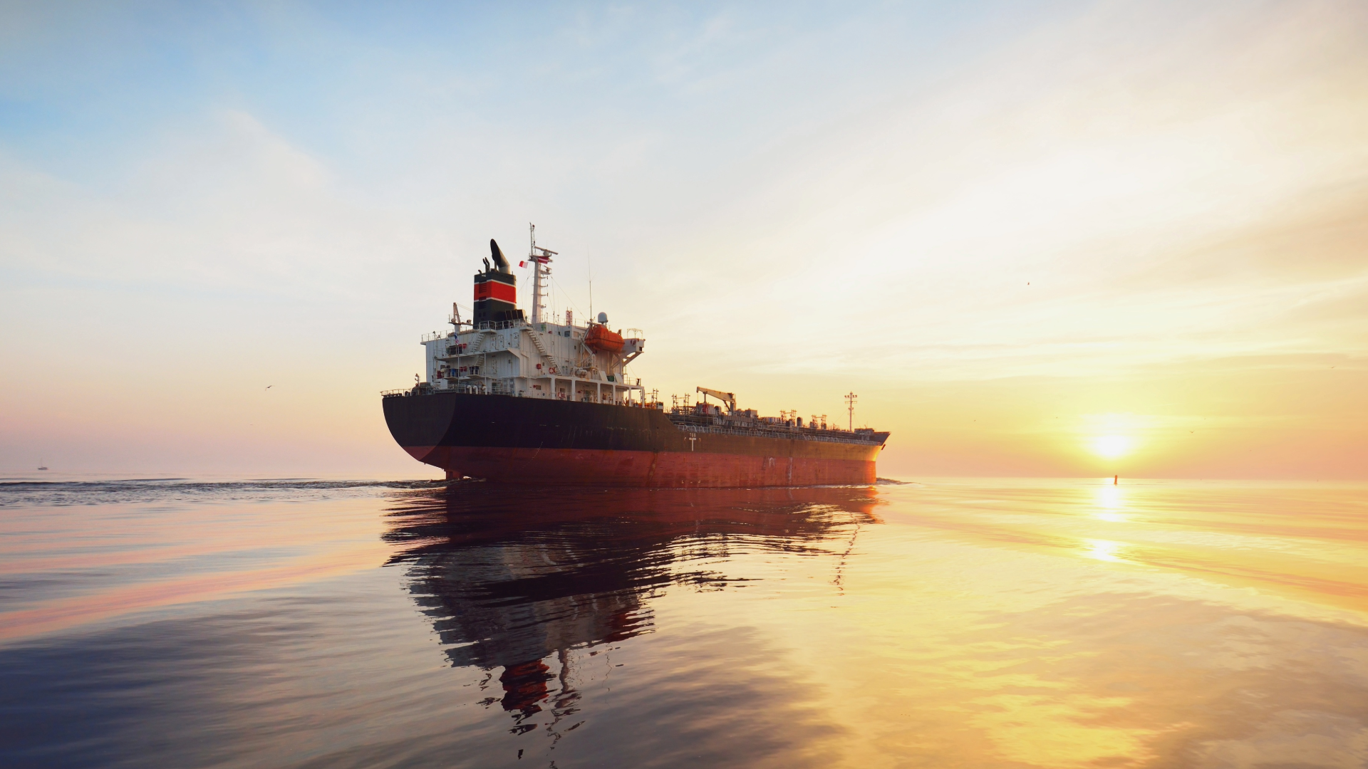 Oil tanker on calm water at sunset, reflecting the colorful sky.