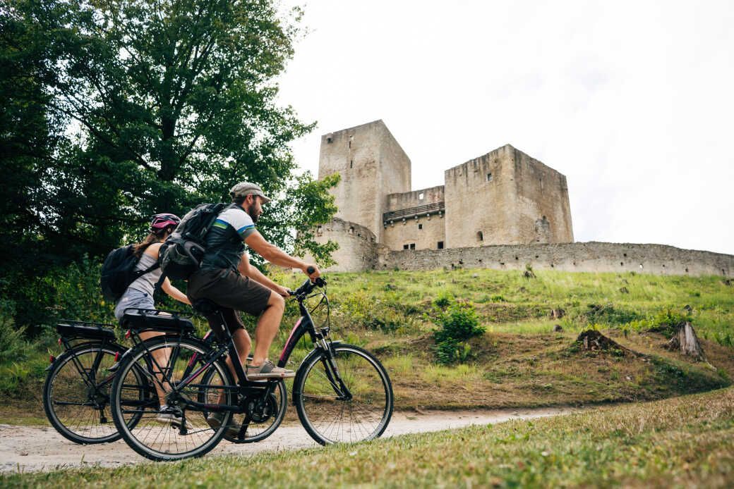 Couple bikes past a medieval castle, sunny day.
