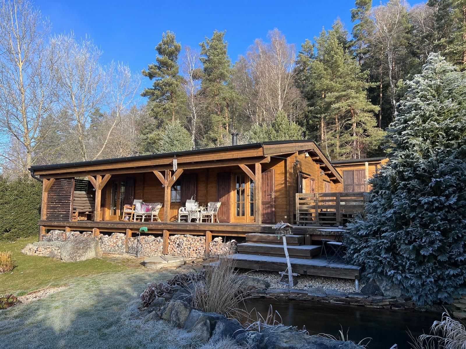 Wooden cabin on a frost-covered hillside, with a porch, chairs, and surrounding trees under a bright blue sky.