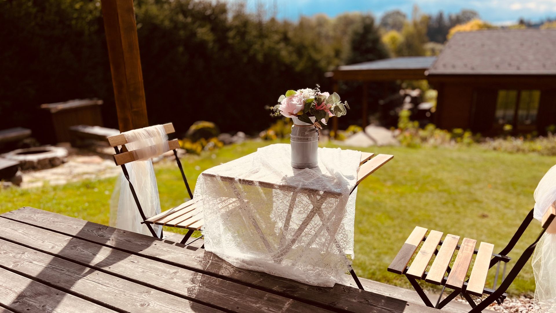 A small table with flowers sits on a wooden deck, flanked by two folding chairs. Backyard setting.