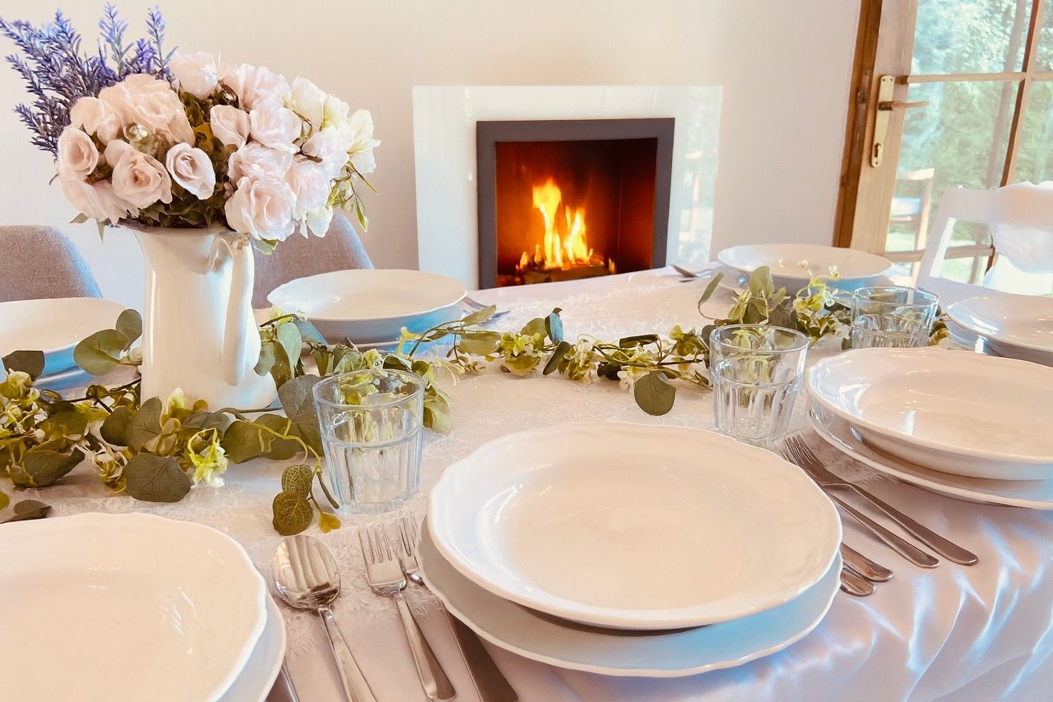 Table set for a meal, flowers in a vase, with a fireplace burning in the background.