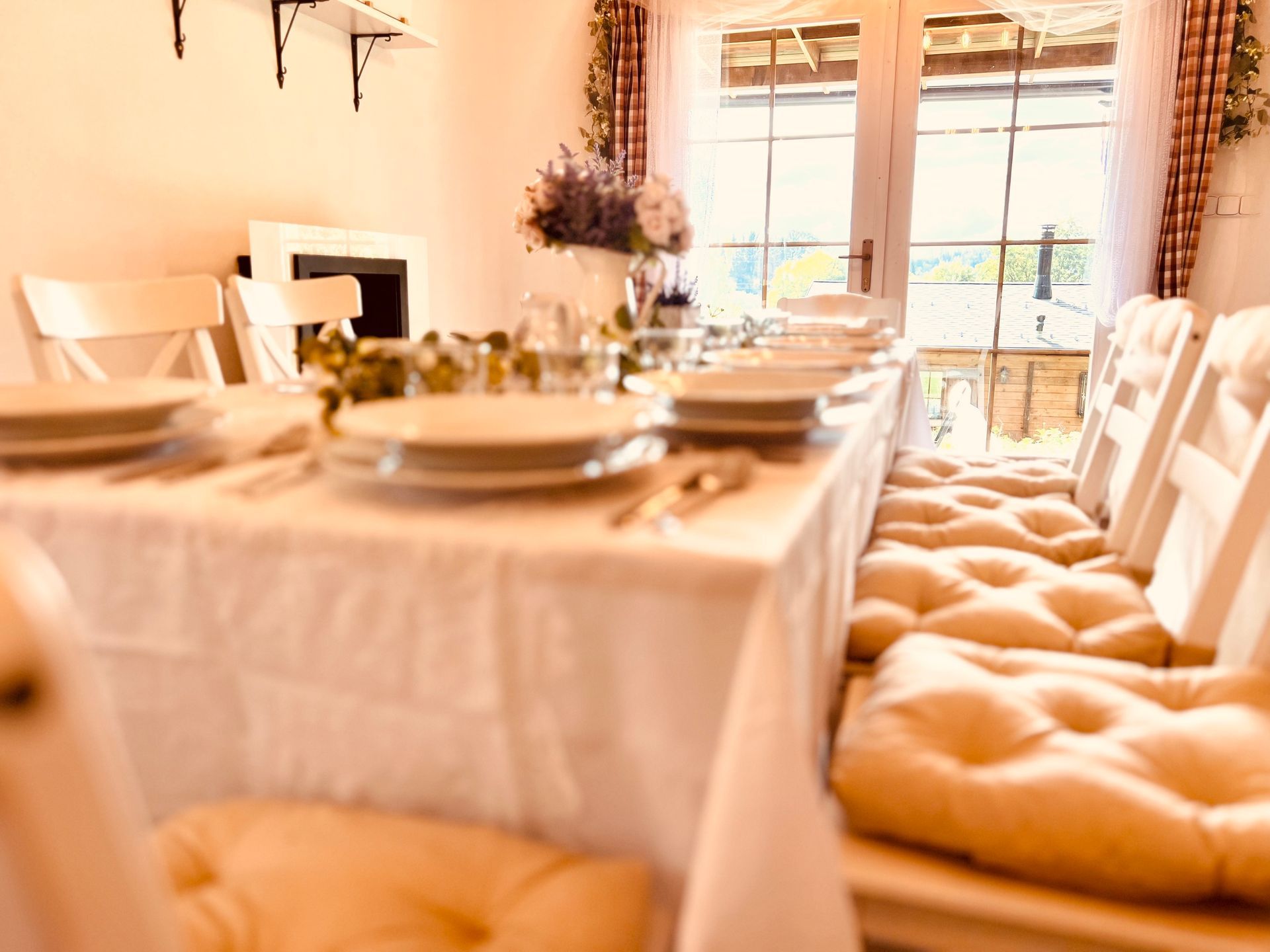Dining table set for a meal with white chairs and cushions, near a window, with flowers on the table.