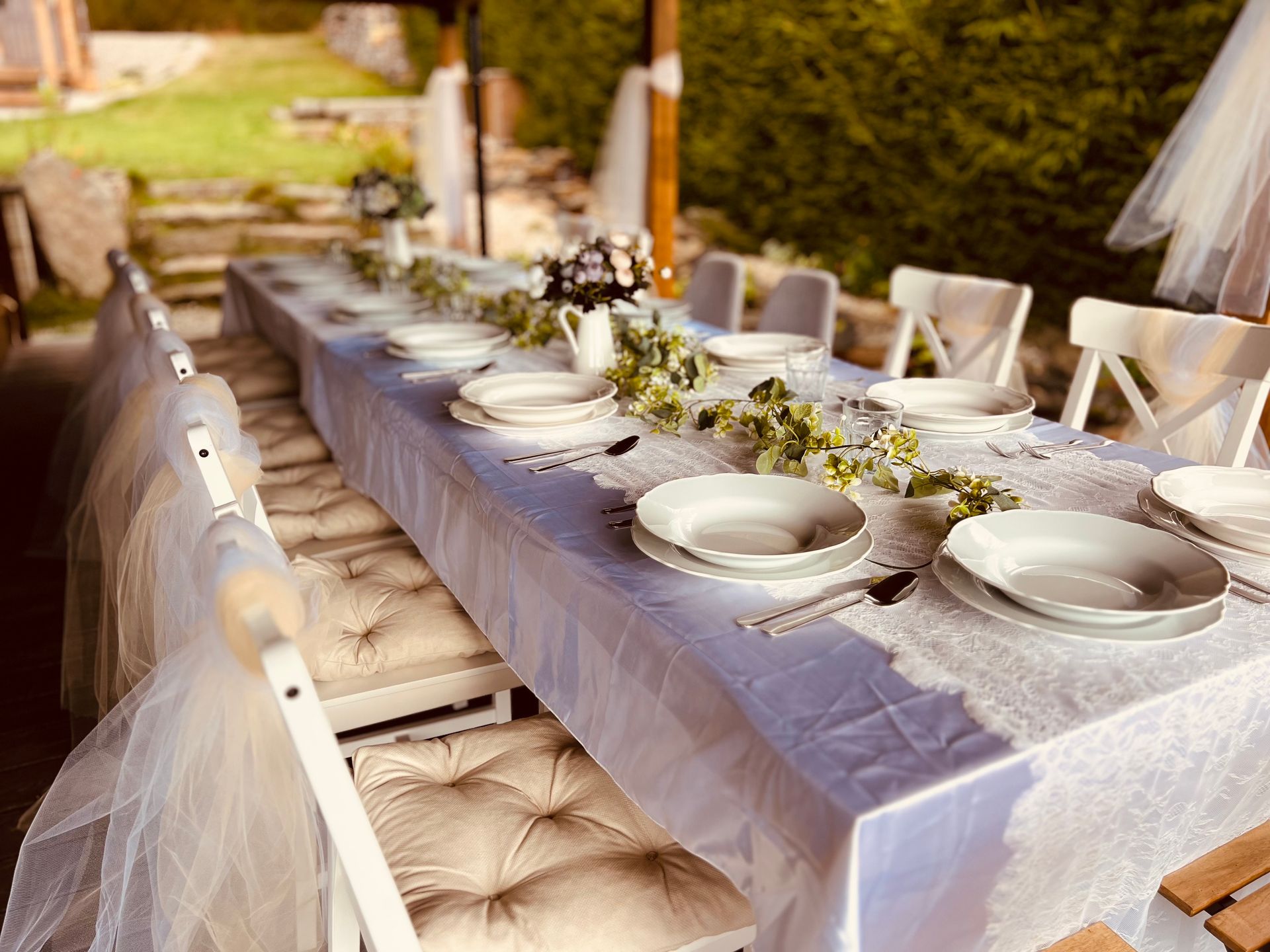 Long dining table set for a celebration outdoors, with white tablecloth, plates, and floral arrangements.