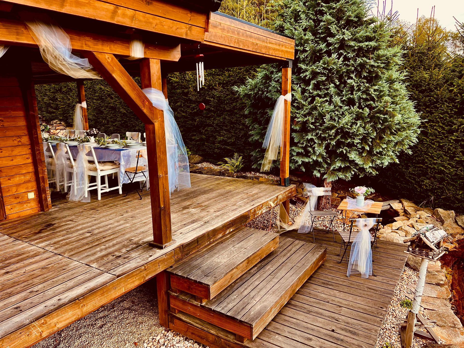 Wooden deck with steps leading to a covered outdoor dining area, surrounded by trees and decorations.