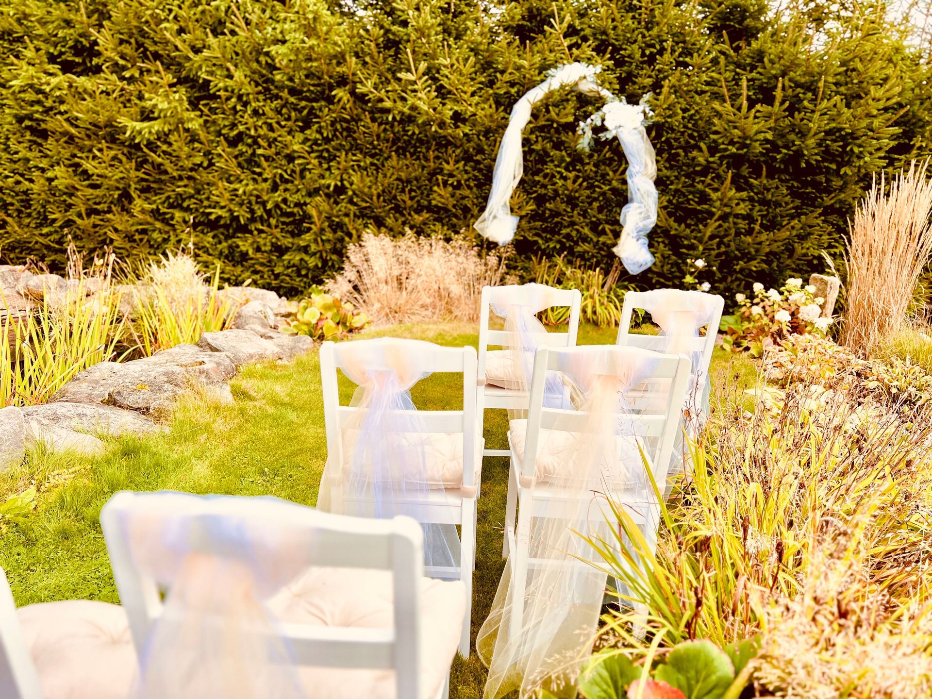 White chairs set up for an outdoor wedding ceremony with a floral arch against a backdrop of greenery.