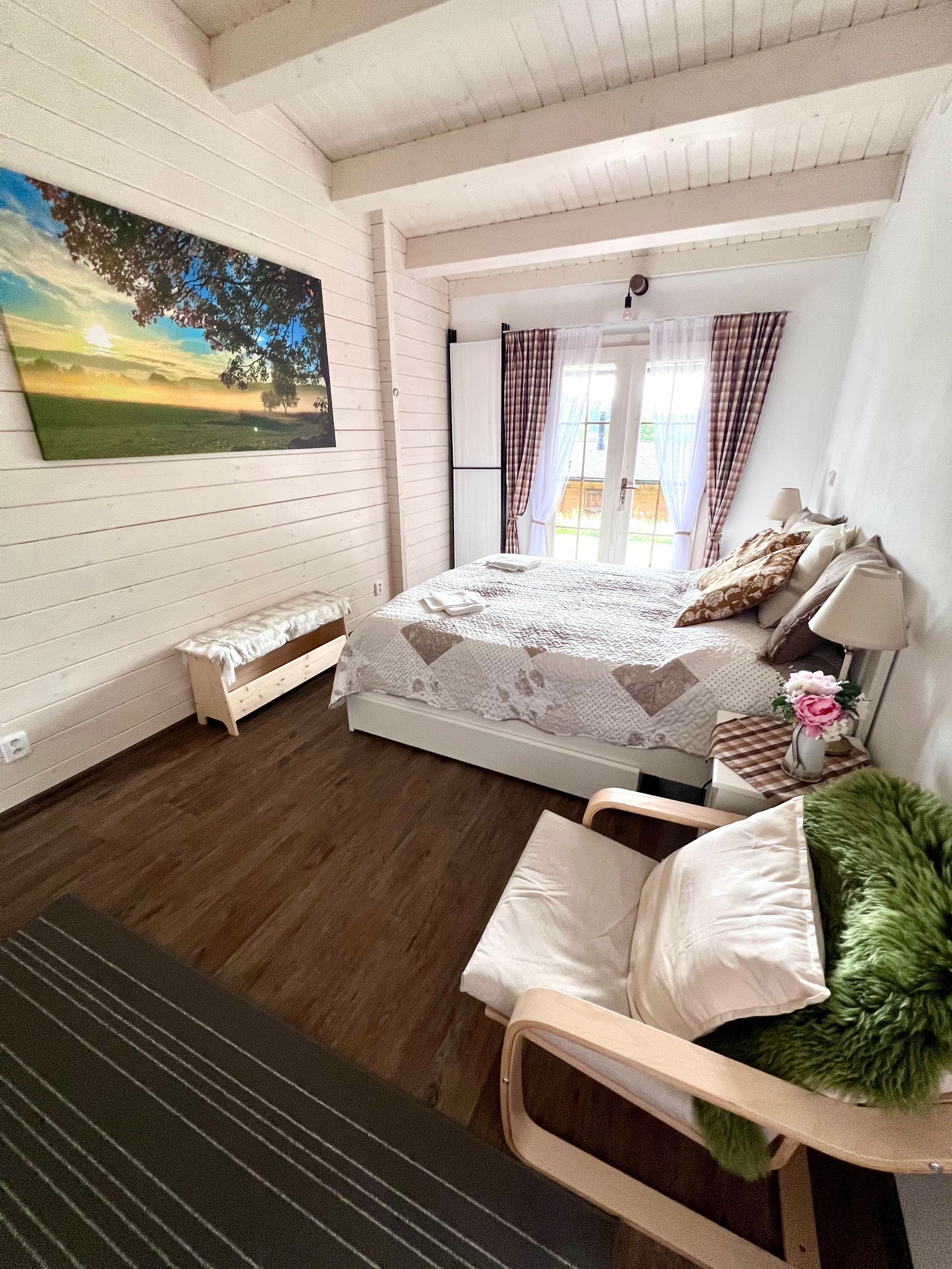 Cozy bedroom with bed, chair, and artwork, wooden floor, white walls and ceiling, and a doorway leading to the outdoors.