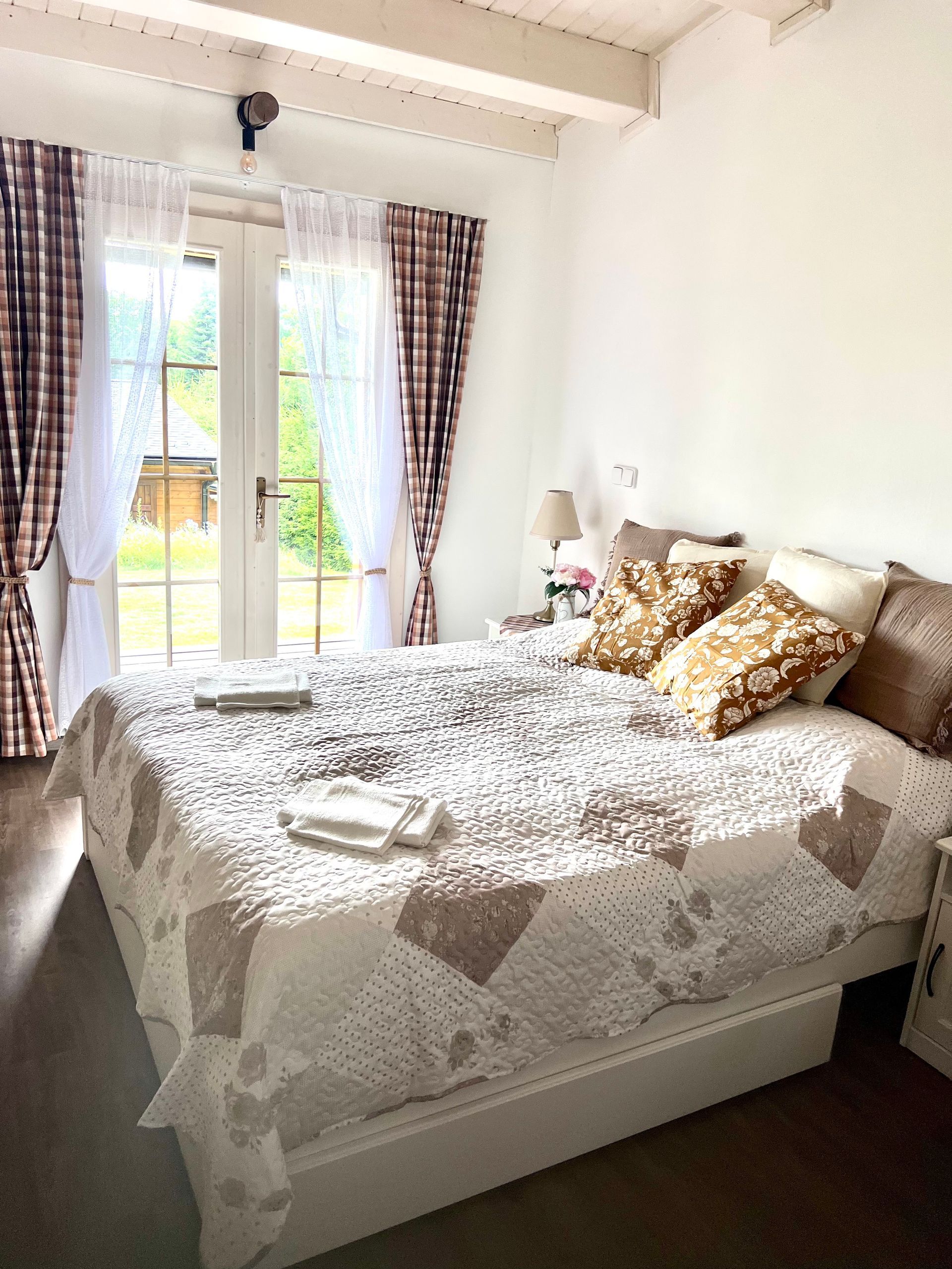 Bedroom with a bed, patterned quilt, and open glass doors overlooking greenery. Neutral tones with brown and white curtains.