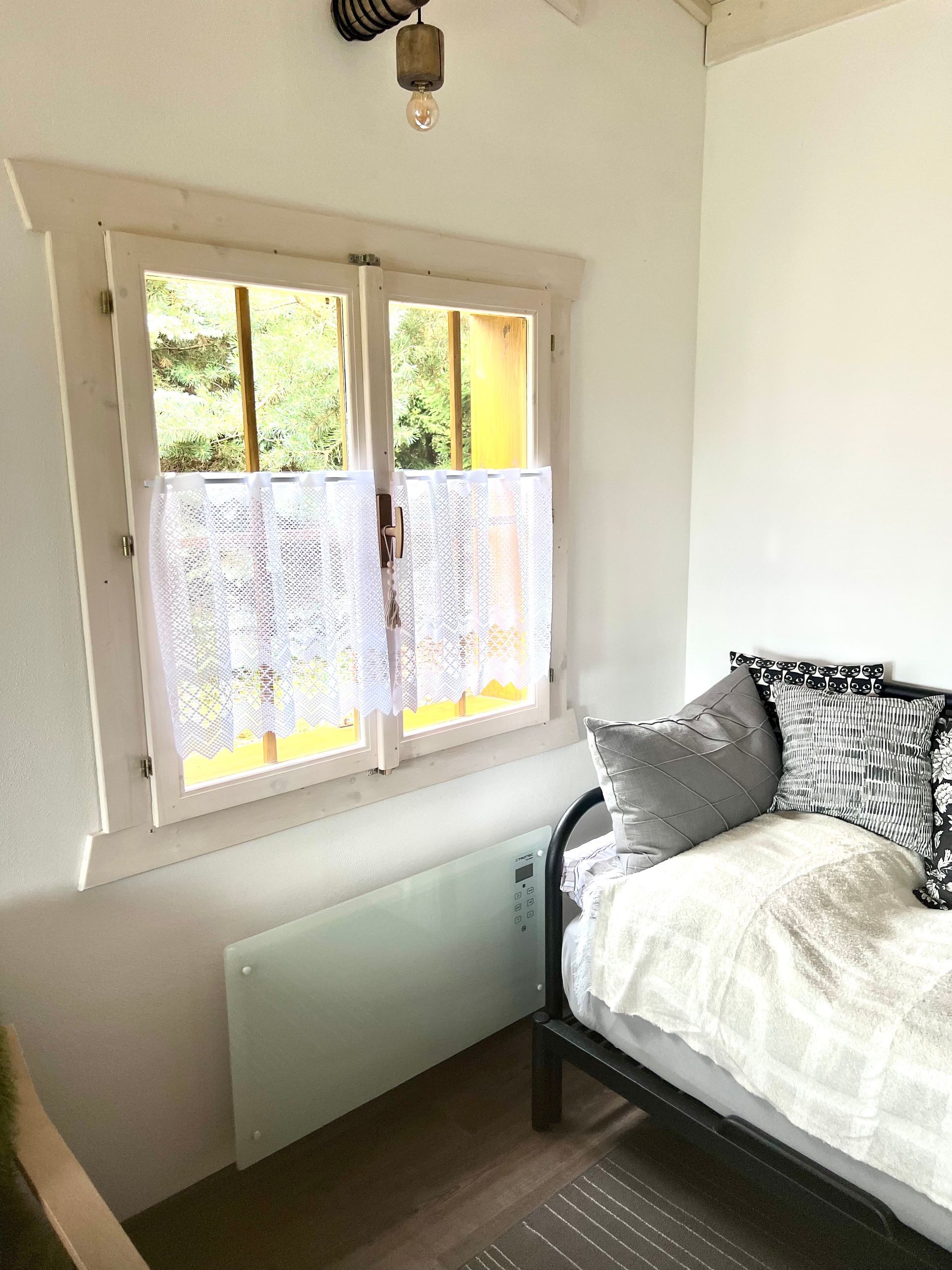 A small room with a window, bed, and a white heater. Sunlight streams in, illuminating the space.
