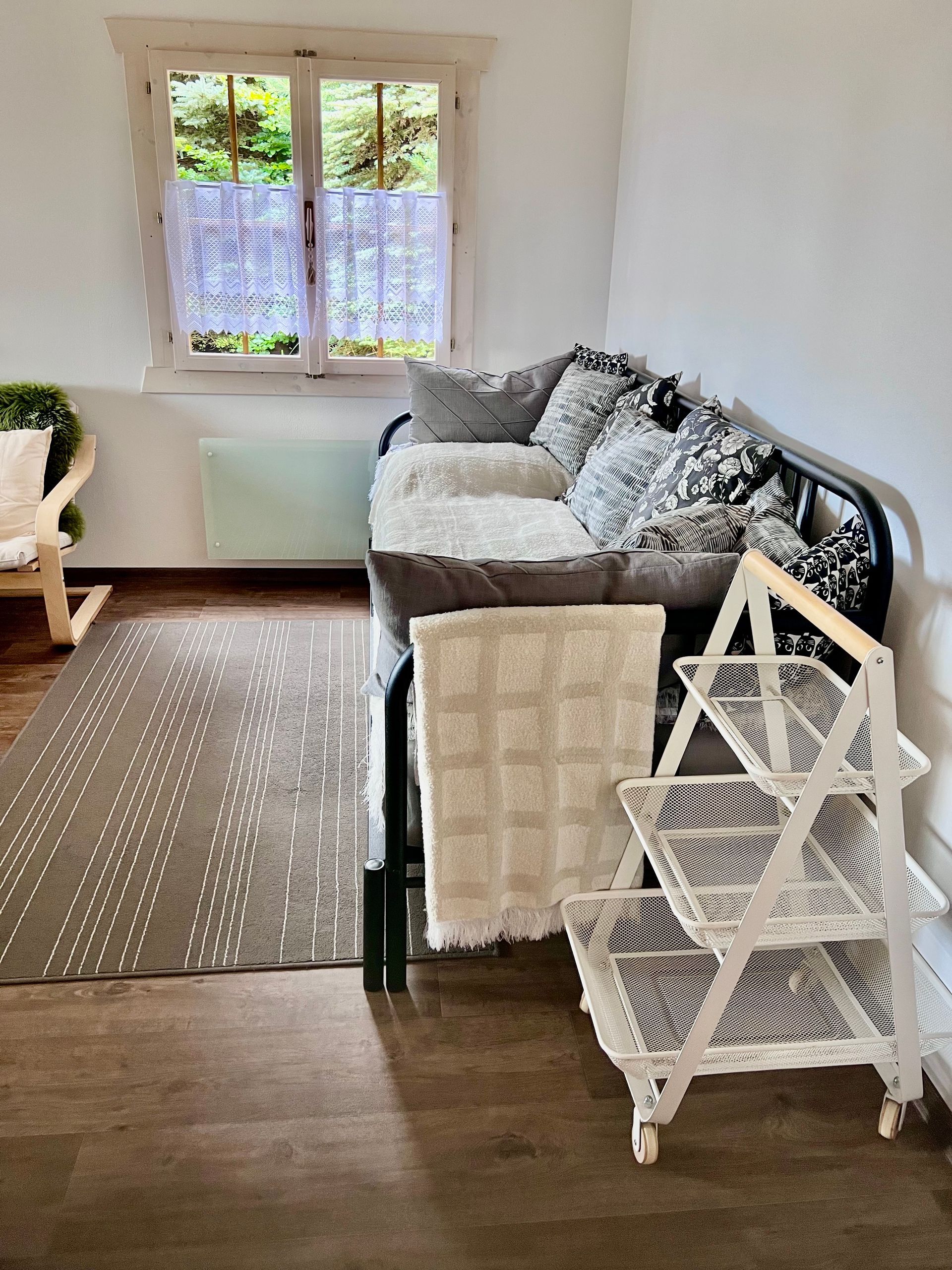 Cozy living area with a daybed, white shelving, and window with sheer curtains. Brown floor with a patterned rug.