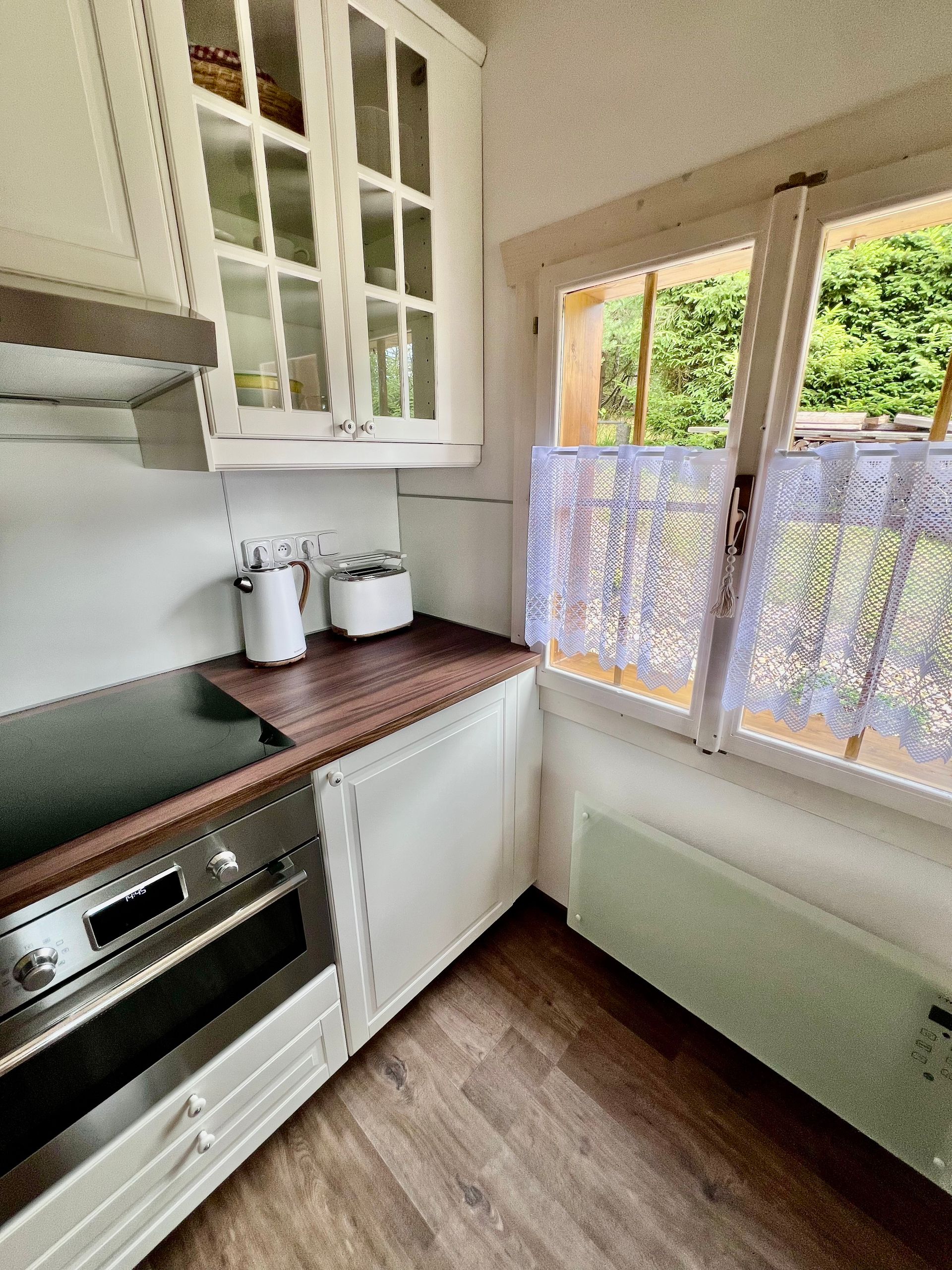 Small white kitchen with cabinets, appliances, wood countertops, and a window with lace curtains.