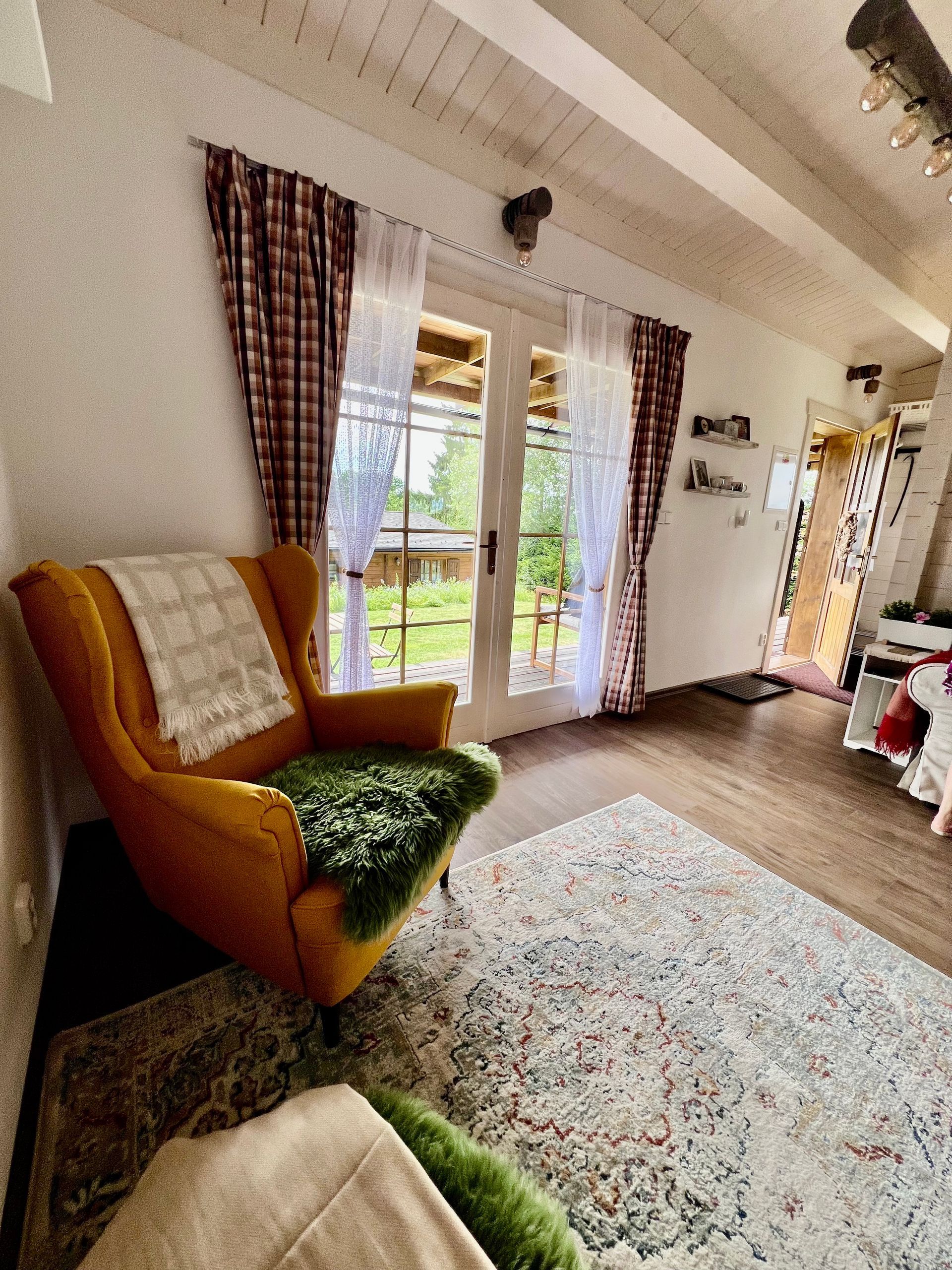 Cozy room with an orange armchair, patterned rug, and French doors leading to a garden. Red plaid curtains.