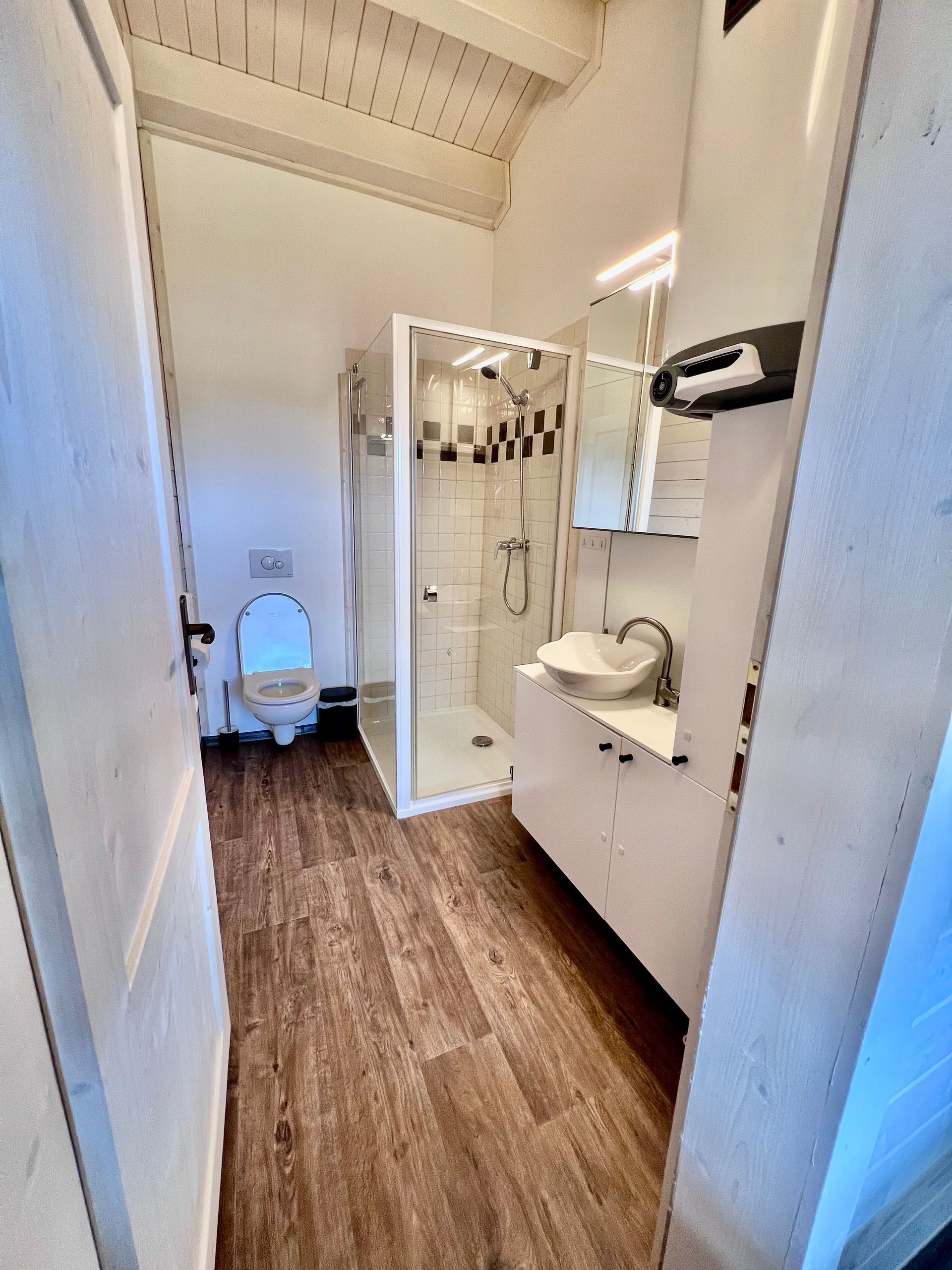 Narrow bathroom with shower, toilet, and sink; wooden floor, white walls.
