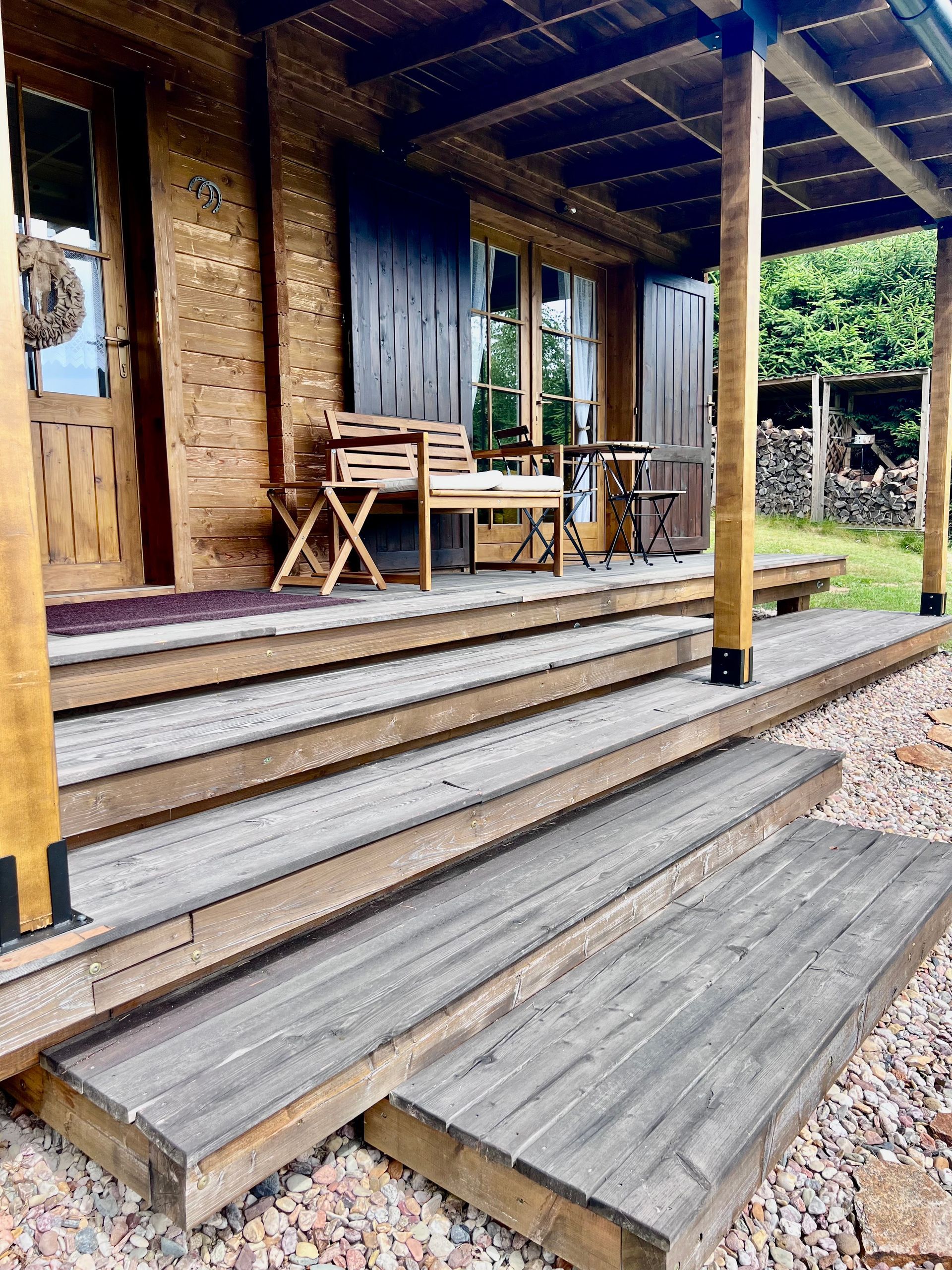Wooden cabin porch with steps; chairs and table; brown wood with grayed deck; gravel ground.