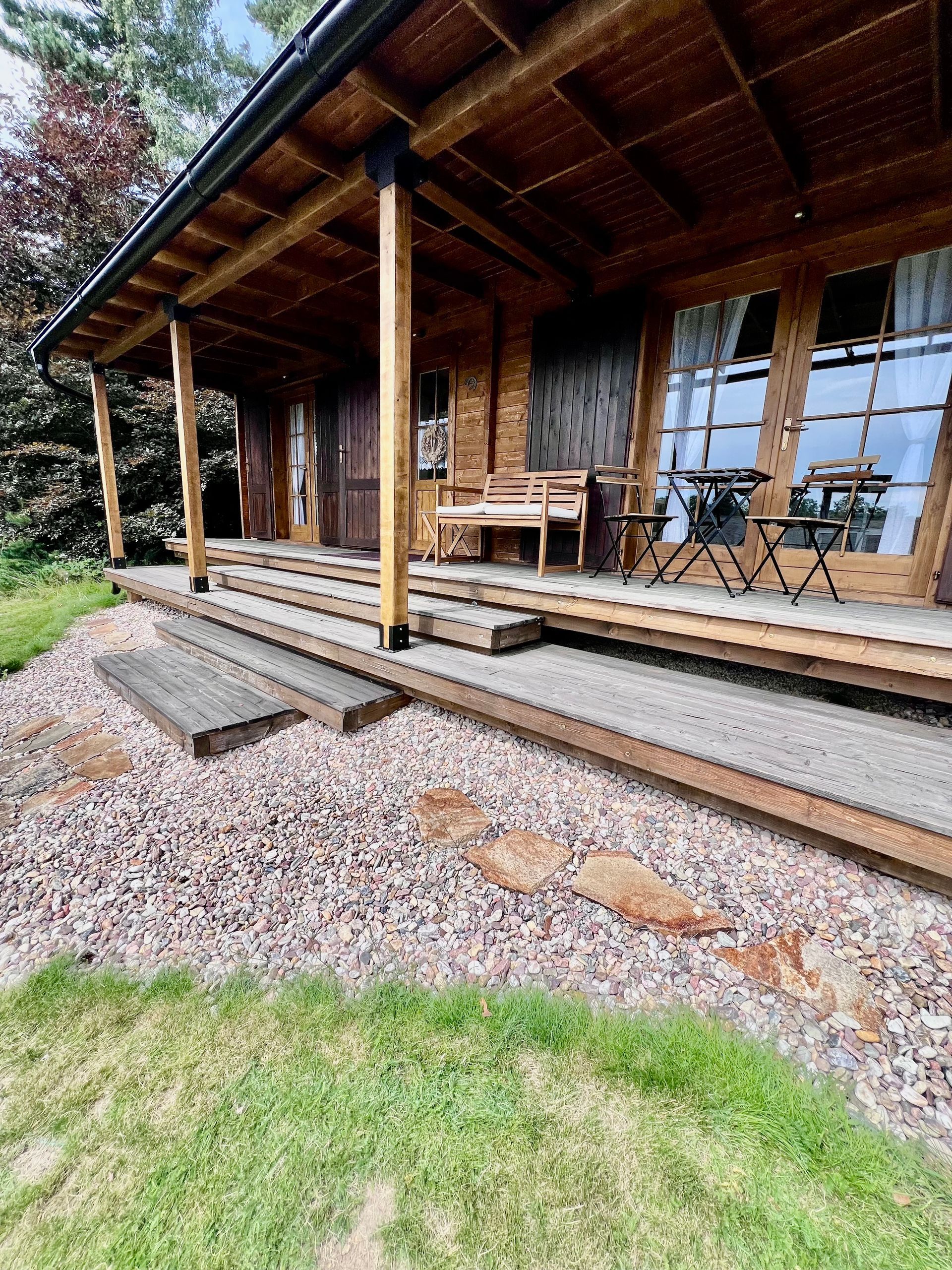 Wooden cabin with porch and gravel walkway, surrounded by grass and trees.