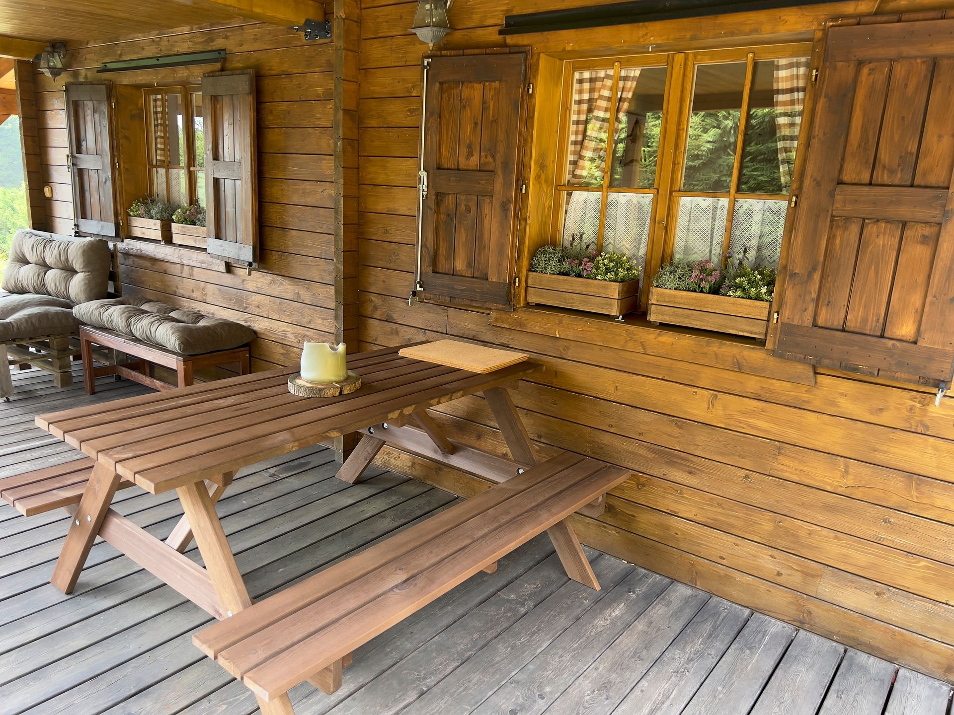 Wooden picnic table and bench on a porch with a log cabin background. Shutters and windows visible.