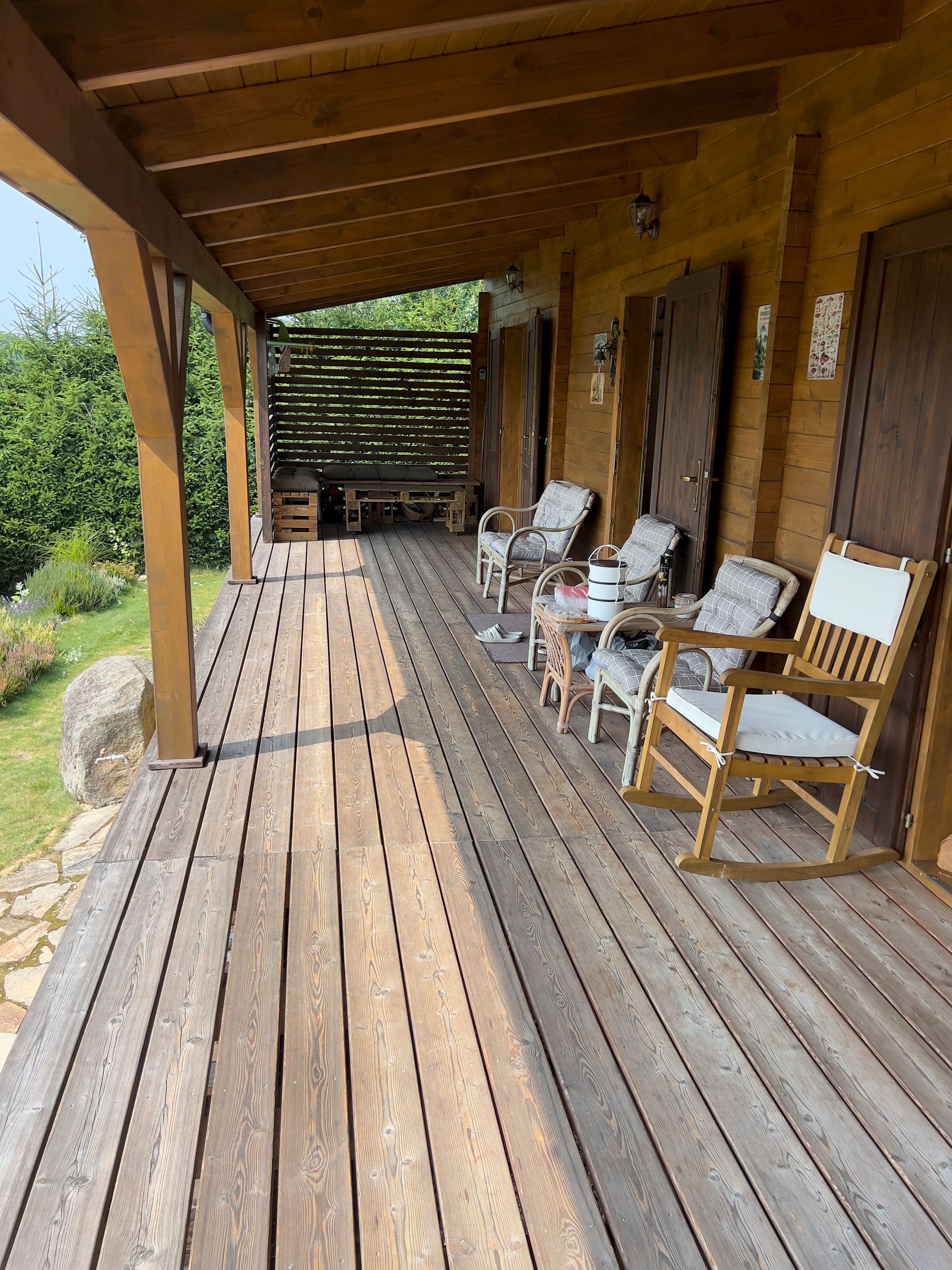 Wooden porch with rocking chairs, overlooking greenery. Brown doors on the right.
