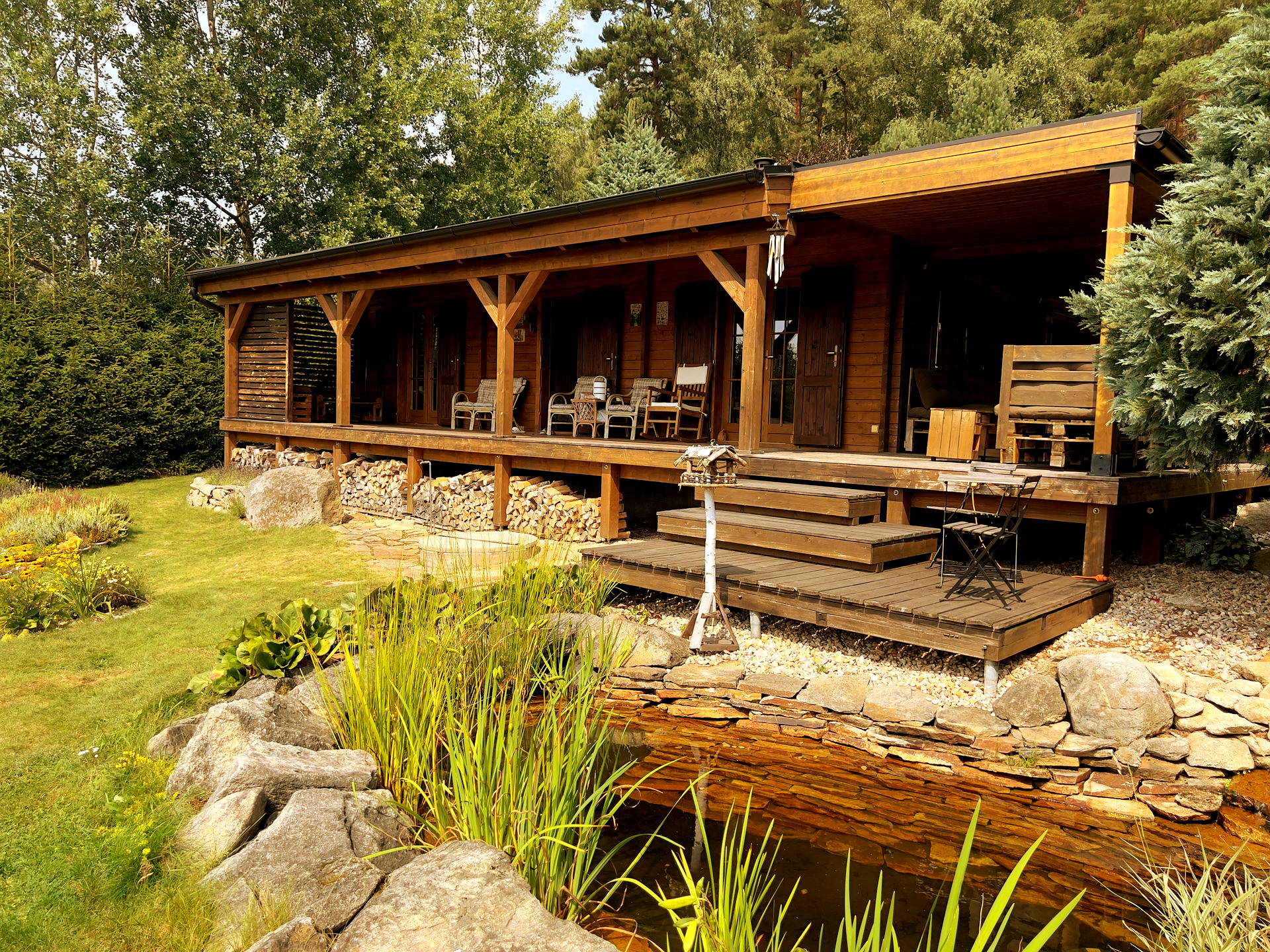Dřevěný srub s verandou s výhledem na jezírko. Dům obklopuje zelená tráva, stromy a kamenné doplňky.