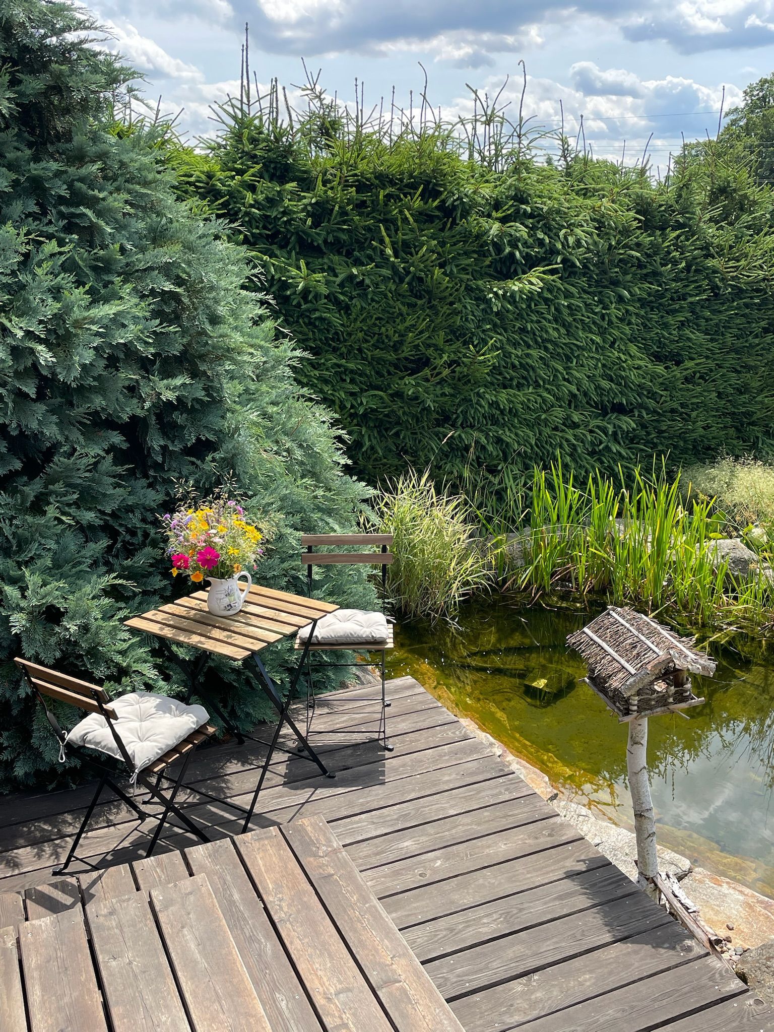 Wooden table and chairs by a pond, surrounded by greenery and flowers; sunny day.