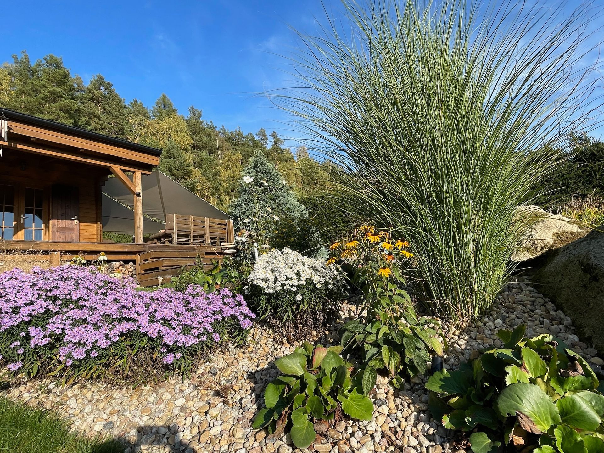 Wooden cabin with flower garden, bright blue sky and trees in the background.