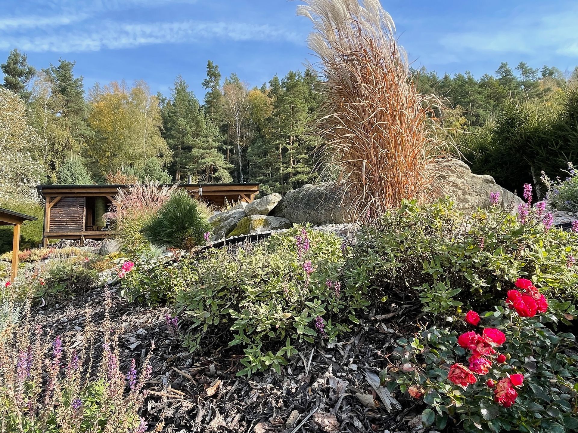 Scenic hillside garden with flowers, shrubs, and a small wooden structure, with trees in the background under a blue sky.