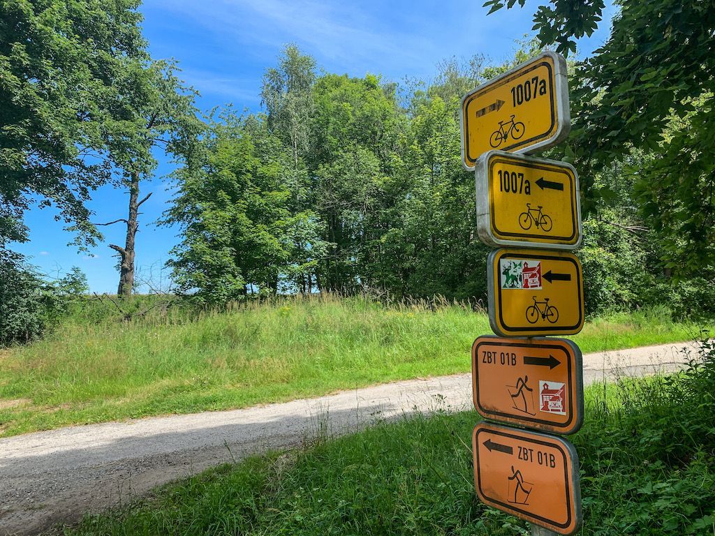 Yellow directional signs on a post along a gravel path indicate bike and hiking trails in a wooded area.